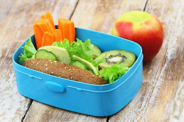 Photo of Healthy school lunch consisting of brown cheese roll, fresh carrots and kiwi fruit and apple