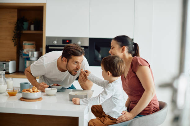 small boy feeding his father while having family breakfast at dining table. - breakfast eating people teens imagens e fotografias de stock