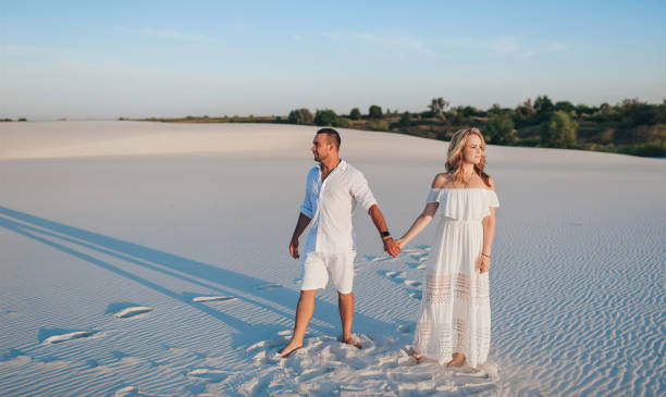 young beautiful couple of a blonde woman and a sunburned guy brunet hold hands. romantic lovers walk on the sand in white clothes at sunset. concept for love, honeymoon, and idyllic relationships. - 5461 imagens e fotografias de stock