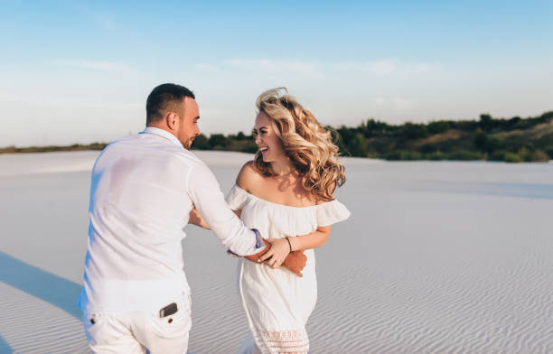 un joven caucásico corre y atrapa y agarra a una hermosa rubia en la arena de la playa. camisa blanca, vestido y pantalones cortos. el concepto de amor, descanso y luna de miel. - honeymoon beach couple heterosexual couple fotografías e imágenes de stock