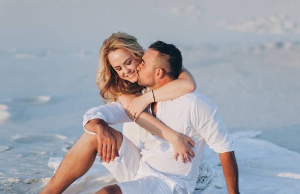 The bearded man and the blonde girl are hugging and kissing while sitting on the white sand and enjoying the sunset. Desert and newlyweds. Love story of cheerful and loving people. Vacation, honeymoon. stock photo