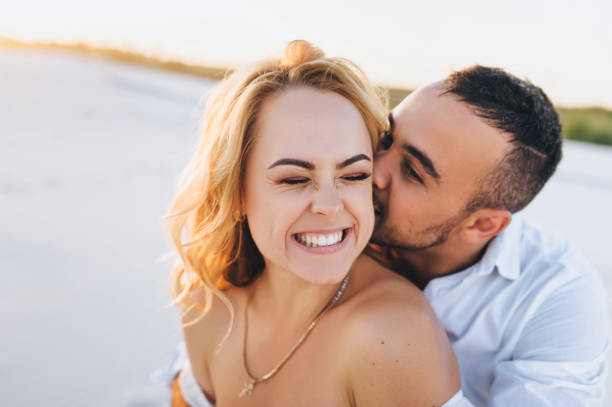 A bearded man and a blond woman hugging against the background of white sand. Portrait of emotional people at sunset. Love in the desert newlyweds. The love story of merry lovers young.Kiss and smile. stock photo