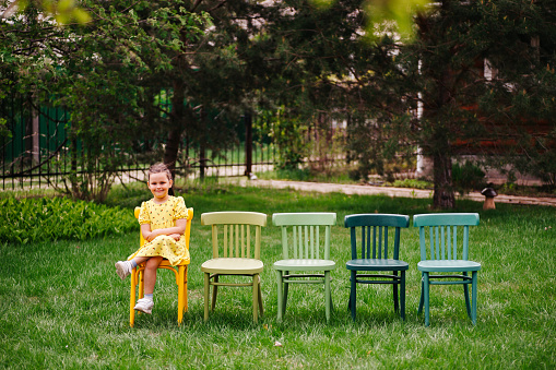a smiling girl in a yellow dress sits cross-legged on Viennese colorful chairs standing in a line in the children's home theater waiting for a play in the park, in summer.