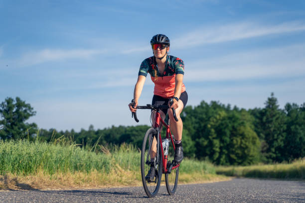 photo en angle, d’une jeune femme cycliste professionnelle, à vélo de route, sur une route goudronnée au milieu de la nature, illuminée par la lumière du soleil. concept d’égalité sportive. - vélo de course photos et images de collection