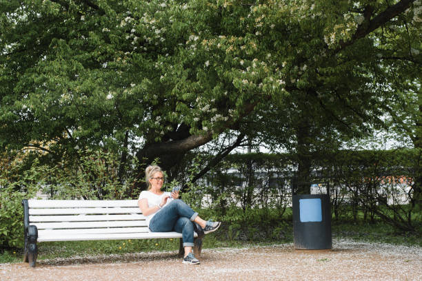 istock Happy woman sitting on a park bench and using a mobile phone outdoors on a summer day. Smiling blonde lady looking at smartphone camera, chatting with someone on social media. Lifestyle summer time 1327994494