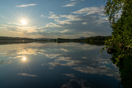Landscape with reflections of sky in a calm lake with forest on the lake shore and a sun star