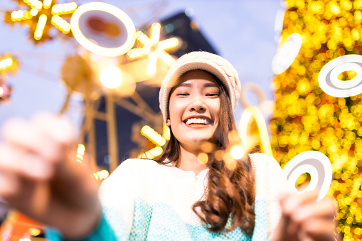 Happy young asian woman taking a selfie photo near christmas tree celebrating New Year. Portrait of young attractive celebrating woman holding sparkles.