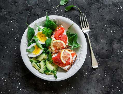 Bowl of green salad, avocado, boiled egg and sandwiches with rye bread, cream cheese and salmon on a dark background, top view. Delicious and healthy breakfast, brunch