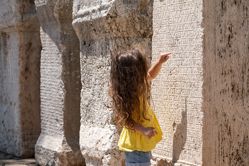 Photo of toddler girl wearing a yellow blouse looking at inscriptions on ancient stone material in public park of Antalya, Turkey. Shot in outdoor with a full frame mirrorless camera.