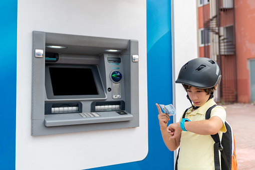 Photo of 8 years old elementary schoolboy wearing a helmet and using smart watch while standing next to ATM. The screen is blank. He is looking at his smart watch and holding 100 Turkish Lira. Shot in outdoor with a full frame mirrorless camera.
