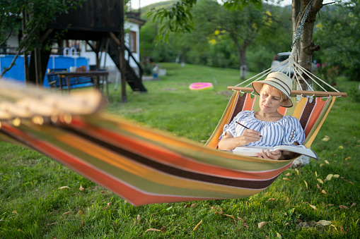 A woman drawing  while resting in a hammock in her backyard
