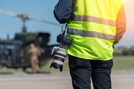 A military photographer, a reporter, in a signal yellow vest, ready to work in front of a military helicopter. Close-up. The background is blurred. High quality photo