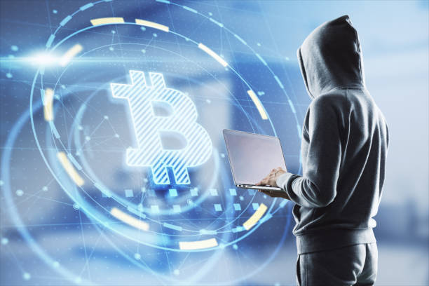 Hacker in hoodie using laptop computer with glowing bitcoin interface in blurry office interior. Hacking, theft and cryptocurrency concept. Double exposure. stock photo