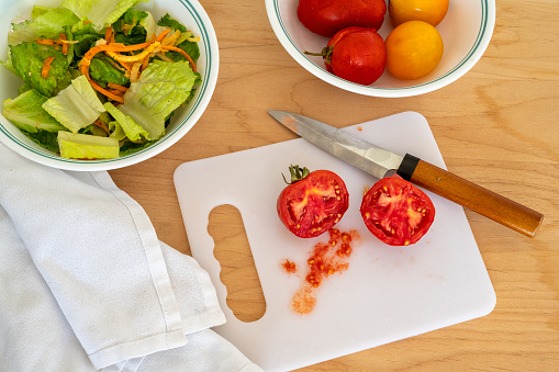 Cherry tomatoes sliced and in a bowl, green salad, knife, cutting board and napkin.