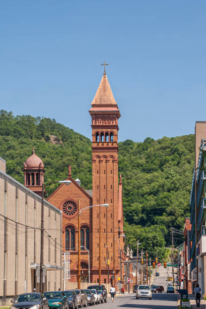 Johnstown, PA, USA - June 11, 2008: Red brick building with 2 towers of Saint John Gualbert Cathedral with Locust Street leading toward belt of green forest on hill under blue sky.
