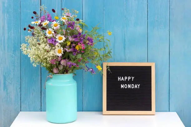 Happy Monday words on black letter board and bouquet of bright wild flowers in tin can vase on table against blue wooden wall. Concept Hello Monday.