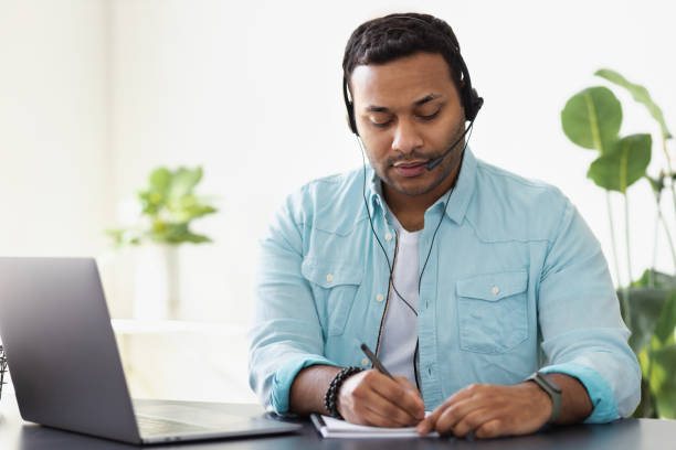 istock Distance work concept. Young indian man support worker or freelancer uses laptop and headset to communicate with the client, makes notes in a notebook, online learning 1328082432