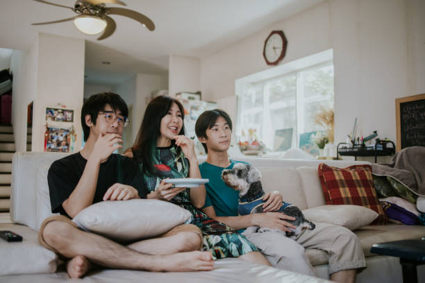 Young Two Asian boys sitting with their mother on sofa in the living room watching TV together on holiday - stock photo Thai Teenage Two boys sitting with their happy mother and lovely dog on the sofa in the living room watching TV together on holiday asian kids watching tv stock pictures, royalty-free photos & images