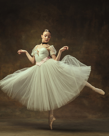 Portrait of a ballerina girl in a white bodysuit, sitting on the stage and tying pointe shoes