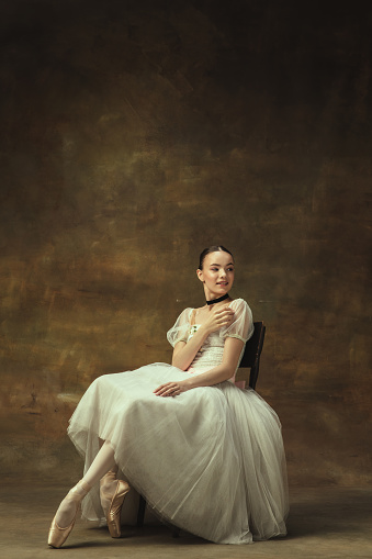 Pretty young woman,, graceful beautiful female ballet dance in white ballet dress sitting isolated on dark studio background. Art, motion, action, inspiration concept. Copy spcae for ad.