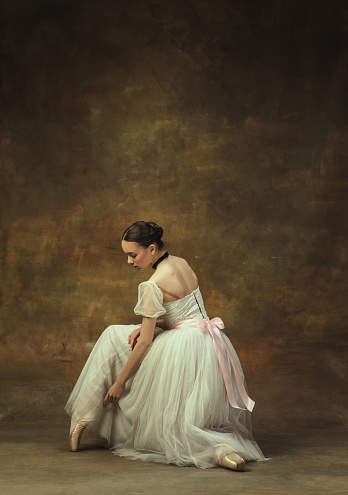 Pretty young woman,, graceful beautiful female ballet dance in white ballet dress sitting isolated on dark studio background. Art, motion, action, inspiration concept. Copy spcae for ad.