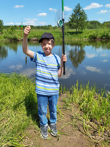 A cute elementary age boy fishes on a river bank with fishing rod. He caught a small fish. A boy is happily showing his catch. Outdoors shooting at sunny summer day at countryside