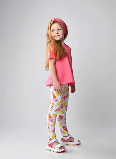 Photo of Red-haired kid girl in summer wear pink t-shirt, colorful pants and sandals stands sideways looking back over shoulder