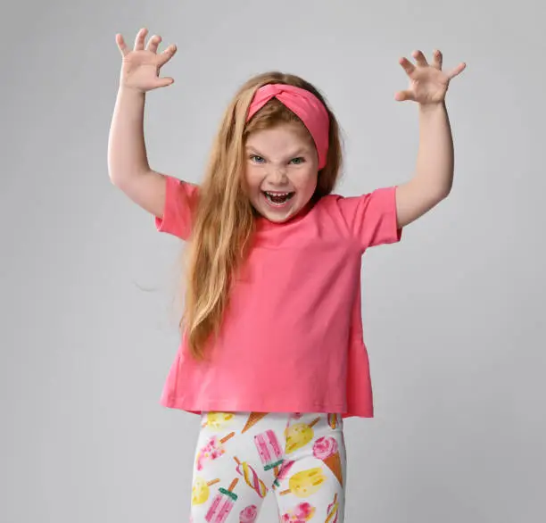 Photo of Playful, frolic redhead kid girl in pink t-shirt and colorful pants plays beast holding hands with claws up