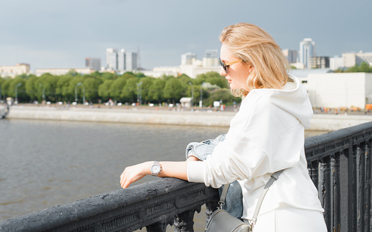 Portrait of woman in sunglasses standing on the bridge and looking at the embankment and river. Young blonde in casual, light clothes on a walk in the city. Travel tourism on a sunny summer day.