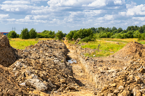Trench for a pipeline with piles of dug soil on the edges against the background of green nature and a sky with clouds