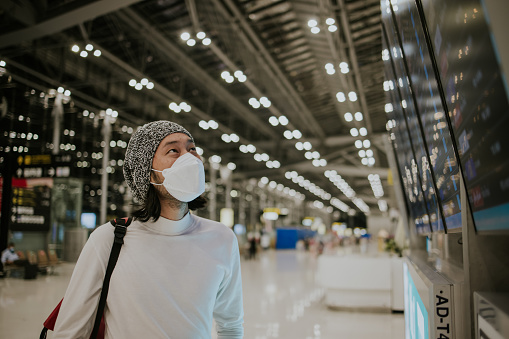 The nightlife of Thai white casual clothing millennial generation male tourist with protective face mask focus on arrival and departure board for checking time of airplane arrival after be vaccinated.