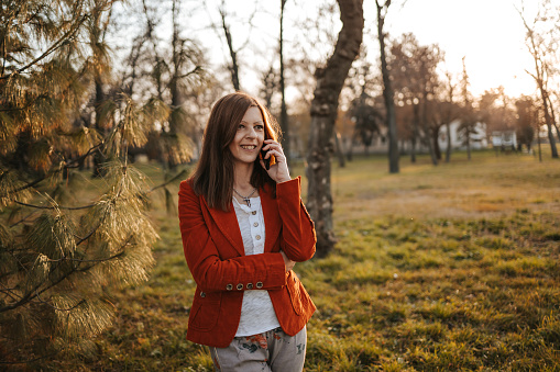 Beautiful young woman in stylish clothes standing in public park in autumn, smiling and talking on smart phone
