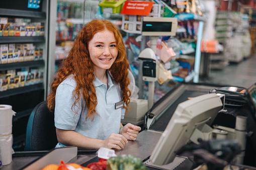 Portrait of a young woman working at grocery store checkout counter. Female cashier sitting behind checkout counter looking at camera and smiling.