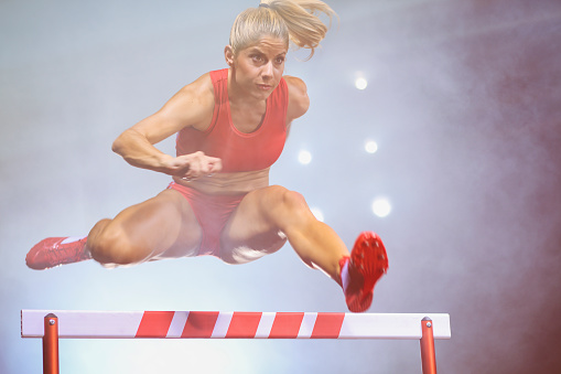 Mid-air shoot of a young woman jumping over hurdle in a track race. Black background.