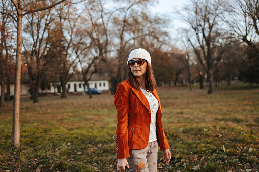 Fashionable beautiful young woman in orange jacket standing in public park in autumn and looking away. Modern city life.