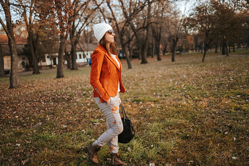 Fashionable beautiful young woman in orange jacket walking with sunglasses and white cap in public park in autumn and looking away. Modern city life.