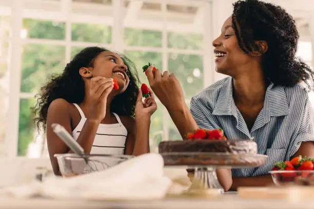 Girl with mother eating strawberries while making make in kitchen. Mother and daughter having fun while baking at home.