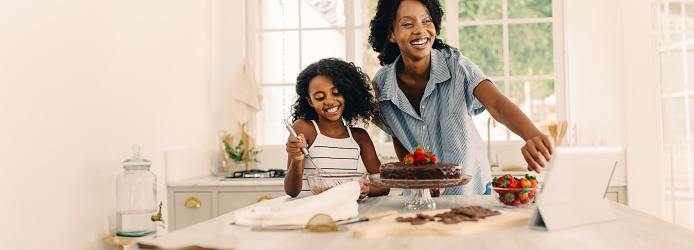 Happy mother and daughter having fun while making cake at home. Woman with girl learning to bake a cake watching online video on a digital tablet in kitchen counter.