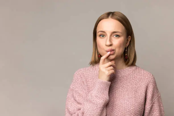 Nervous young female biting finger nail thinking making doubtful decision or choice stand wear pastel pink sweater isolated over gray studio wall. Confused caucasian blond woman millennial wondering