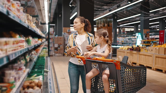 Woman takes packed products from the shelf, packed mushrooms and puts them in a trolley. Little girl and her mom, housewife, enjoy family shopping together