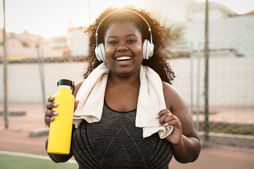 Feliz mujer africana con curvas haciendo ejercicios y rutina de ejercicios mientras escucha música con auriculares inalámbricos al aire libre photo