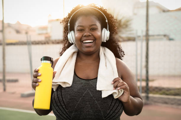 Happy curvy African woman doing jogging and workout routine while listening music with wireless headphones outdoor stock photo