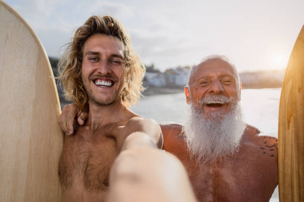 happy fit surfer machen selfie und spaßigen gemeinsam bei sonnenuntergang - extremsport lifestyle und freundschaftskonzept - surfen fotos stock-fotos und bilder