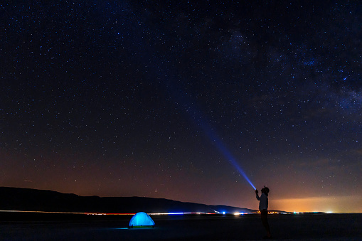 yıldızlar altında kamp yapan adam, arkada şehir ışıkları ve yoldan geçen araba ışıklarının izlei görülmektedir. gece karanlığında uzun pozlanmış fotoğraf, full frame makine ile çekilmiştir.