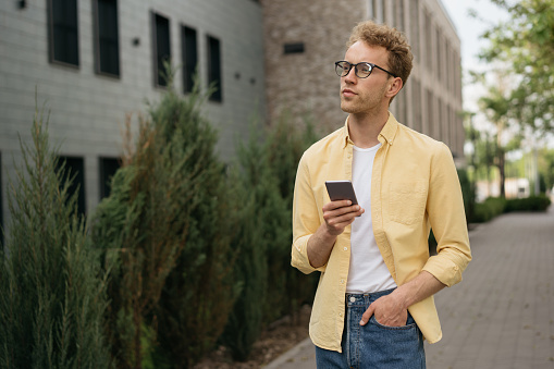Handsome pensive man wearing yellow casual shirt and stylish eyeglasses using mobile phone walking on the street, copy space