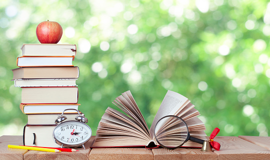 Open book with magnifying glass, bell, silver watch, colours pencils, stack of books and red apple on the nature blurred background with bokeh lights