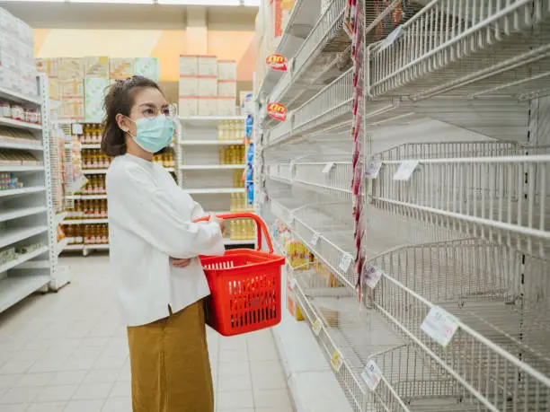 Asian woman looking at many empty shelves with few canned goods left in a Bangkok supermarket because of panic shopping because of Covid-19 virus. Thailand.