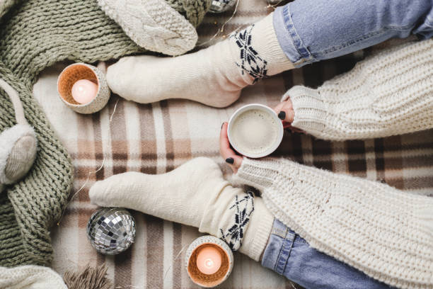 Young woman sits on plaid in cozy knitted woolen white sweater and socks holds cup of cocoa in her hands. Hygge New Year, cozy Christmas, preparation for holidays. Candles, Christmas balls. stock photo