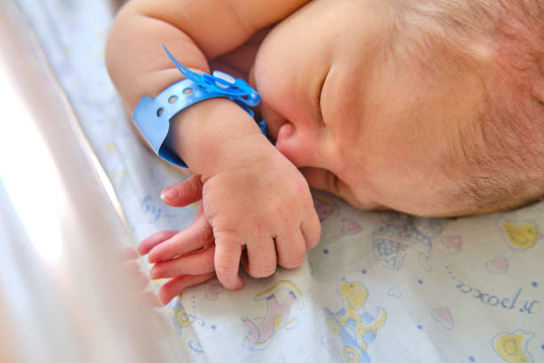 A newborn baby with a maternity hospital bracelet on his arm is sleeping in a crib. A newly born child in a clinic bed behind a transparent glass A newborn baby with a maternity hospital bracelet on his arm is sleeping in a crib. A newly born child in a clinic bed behind a transparent glass baby bracelet stock pictures, royalty-free photos & images