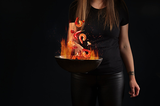 Closeup of peeled shrimp tails, hot pepper, tomatoes and garlic hovering in fire over wok pan in hands of young woman on black background. Female chef throwing ingredients into air. Cooking concept